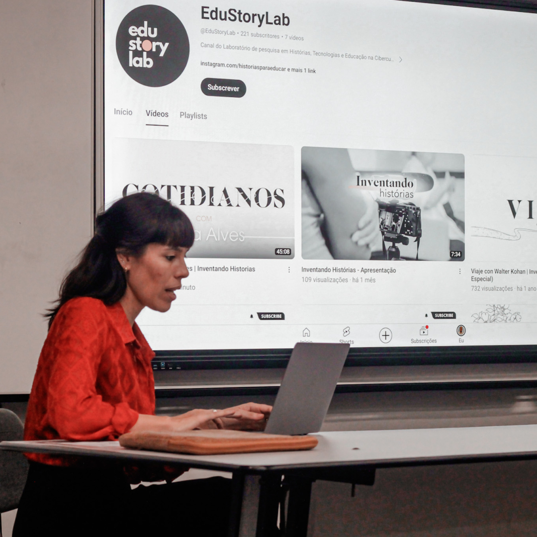 Tania sentada com computador, na sala da UERJ, no dia do lançamento do episódio "Cotidianos, com Nilda Alves".